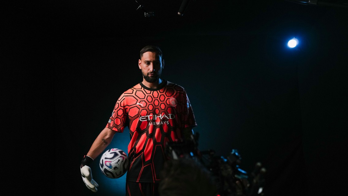 A soccer player wearing the Manchester City goalkeeper kit, holding a soccer ball, with Etihad Airways logo visible, in a dimly lit setting.