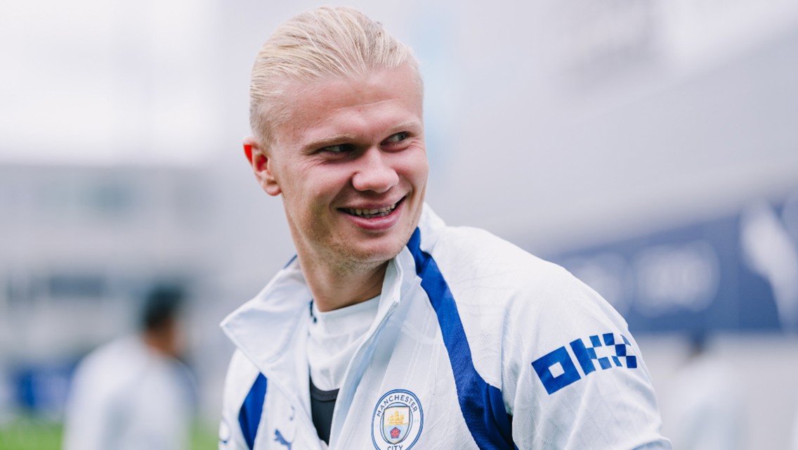 A man wearing a Manchester City training jacket featuring the club's logo, smiling in an outdoor setting.