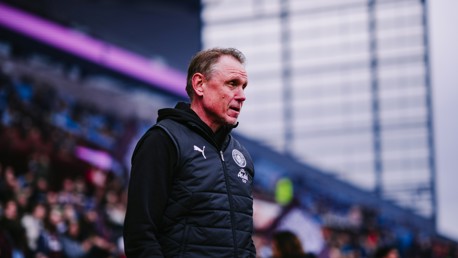 A person in a black sports jacket stands by the sidelines of a football stadium with blurred crowd and architecture in the background.
