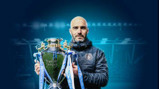 A person wearing a Manchester City jacket is holding a large trophy with blue ribbons in a stadium setting.