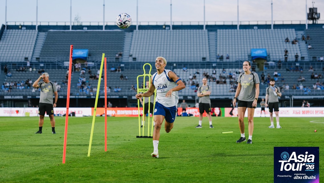 A soccer player from Manchester City is training on the field with coaching staff present during the Asia Tour. The stadium seats are partially filled.