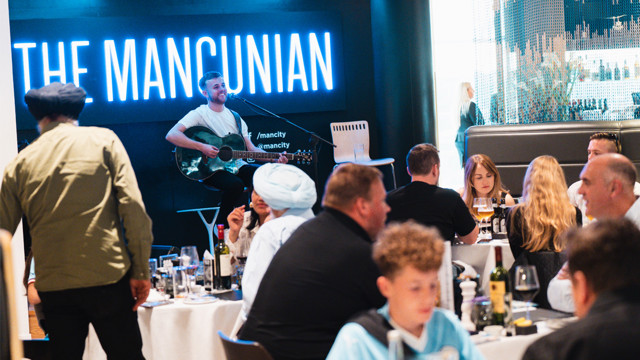 A musician performs with a guitar at a dining event in a venue named 'The Mancunian.' Guests are seated at tables enjoying food and drinks.