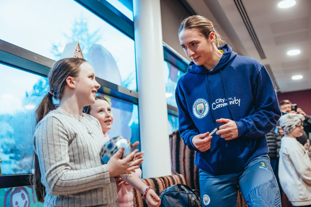 Manchester City player engages with a young fan, wearing a 'City in the Community' hoodie, indicating CITC involvement.