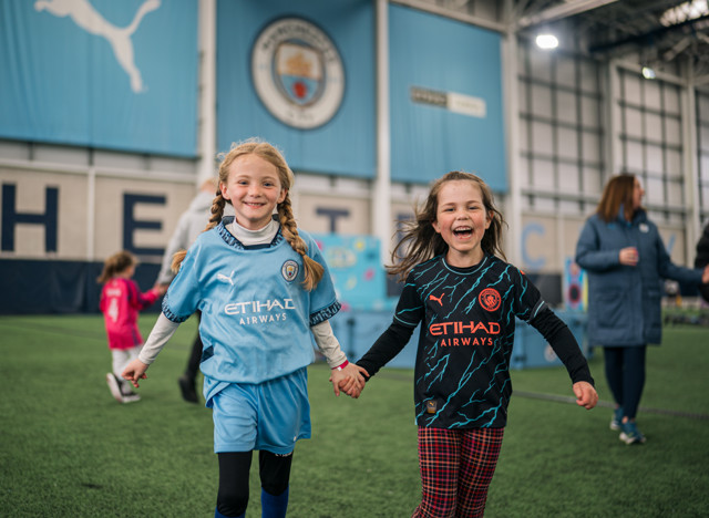 Two young girls holding hands, wearing Manchester City football kits, inside a sports facility with Man City logos and branding visible in the background.