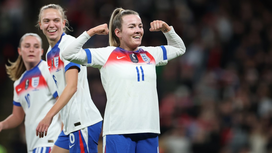 An England women's football team player celebrating by flexing her arms. Two teammates are partially visible with blurred faces, wearing white jerseys with blue numbers and logos.