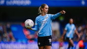 A Manchester City women's soccer player wearing the team's uniform holds a soccer ball and gestures on the field during a match.