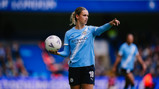 A Manchester City women's soccer player wearing the team's uniform holds a soccer ball and gestures on the field during a match.