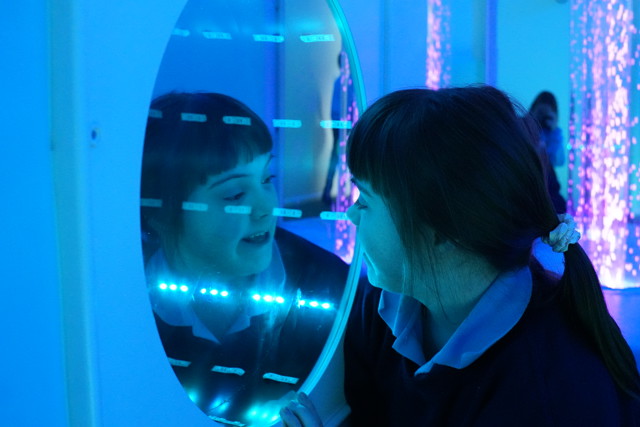 A child with long hair wearing a scrunchie is looking into an interactive mirror in a sensory room with blue lighting.