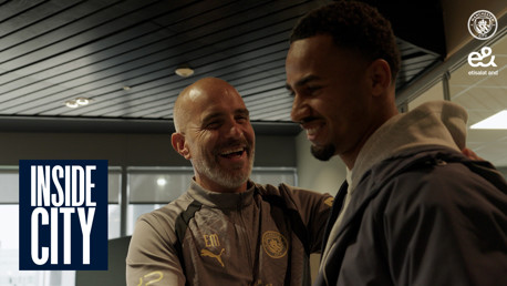 Two individuals smiling and interacting in a setting with a Manchester City logo and 'Inside City' text visible. One wears a jacket, the other is in Manchester City sportswear.