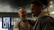 Two individuals smiling and interacting in a setting with a Manchester City logo and 'Inside City' text visible. One wears a jacket, the other is in Manchester City sportswear.