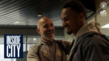 Two individuals smiling and interacting in a setting with a Manchester City logo and 'Inside City' text visible. One wears a jacket, the other is in Manchester City sportswear.