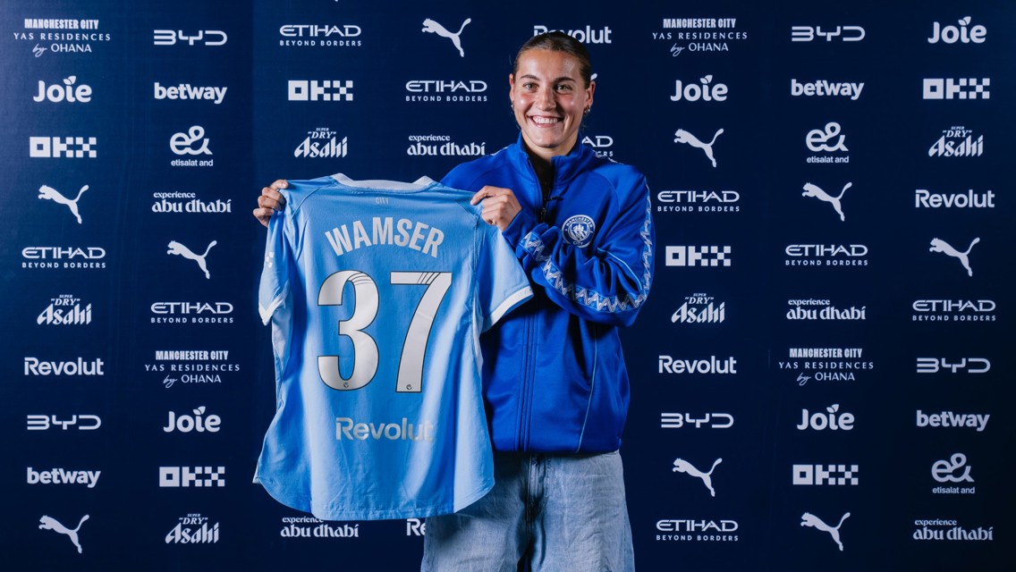 A person smiling while holding a Manchester City jersey with the name 'Wamser' and number '37'. The background features sponsor logos including Puma, Etihad, and Revolut.