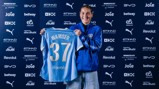 A person smiling while holding a Manchester City jersey with the name 'Wamser' and number '37'. The background features sponsor logos including Puma, Etihad, and Revolut.