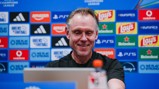 A man smiling at a computer during a press conference, with a backdrop featuring various sponsor logos such as Amazon, Adidas, Lay's, and Heineken.