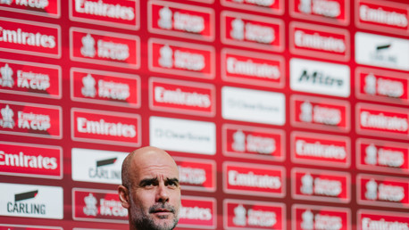 A person with a blurred face at a press conference with a red Emirates FA Cup branded background featuring brand logos.