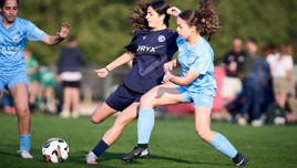 Two girls are playing soccer on a field, wearing blue and navy uniforms. One girl in navy is attempting to play the ball, while the girl in blue tackles her.