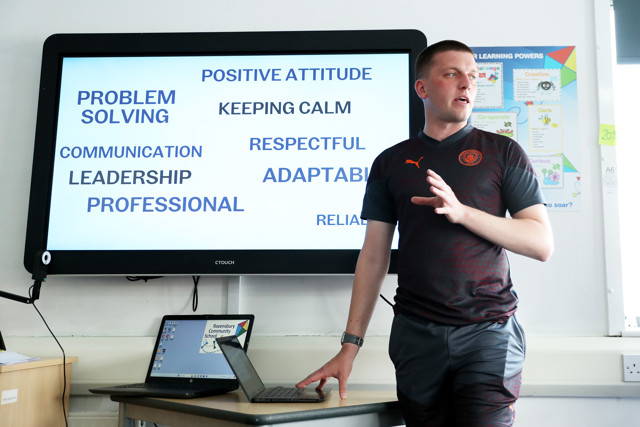 A person giving a presentation on team skills with a screen displaying words such as PROBLEM SOLVING, COMMUNICATION, LEADERSHIP, PROFESSIONAL, POSITIVE ATTITUDE, KEEPING CALM, RESPECTFUL, ADAPTABLE, RELIABLE.