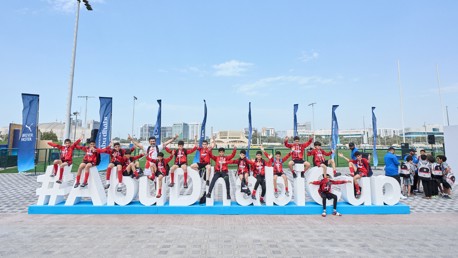 A group of individuals is sitting and posing on a large hashtag sign that reads '#AbuDhabiCup' outdoors. Several banners with logos and text are visible, suggesting a sports event setting.