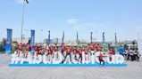 A group of individuals is sitting and posing on a large hashtag sign that reads '#AbuDhabiCup' outdoors. Several banners with logos and text are visible, suggesting a sports event setting.