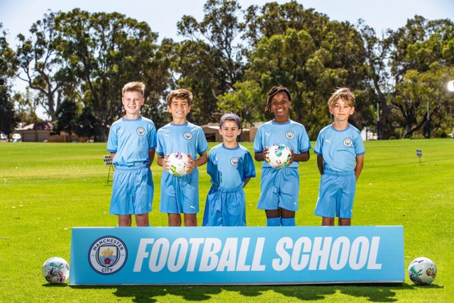 Five kids in Manchester City youth football uniforms stand in front of a sign that reads 'FOOTBALL SCHOOL' on a grassy field. They are holding painted soccer balls.