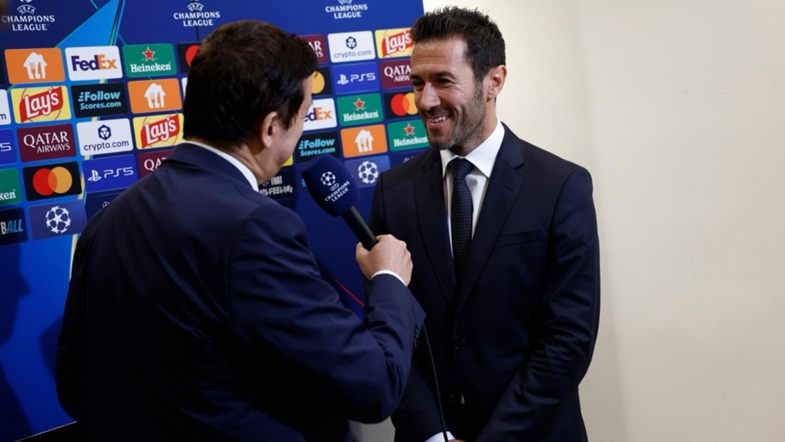 Two men in suits during an interview with a UEFA Champions League backdrop featuring various sponsor logos.