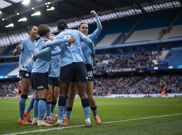 A group of football players wearing light blue jerseys celebrating a goal at the Etihad Stadium. The players are hugging and raising their arms in celebration, with a stadium crowd in the background.