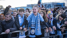 A crowd of football fans wearing Manchester City scarves and clothing, gathered behind a barrier, showing excitement and support.