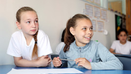 Two children are sitting at a classroom table. One is holding a pencil, and there is a white sheet of paper on the table. Both children have their faces blurred.