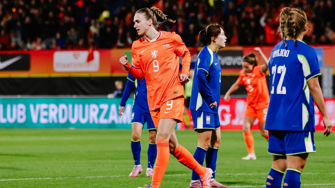 A female football player in an orange jersey with number 9 celebrates a goal. She is surrounded by players in blue jerseys. The background includes a blurred crowd and advertisements.