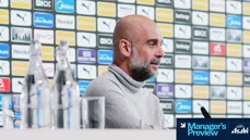 A manager sits at a press conference table before a backdrop featuring multiple logos, including Puma and Etihad, with water bottles and a microphone visible.