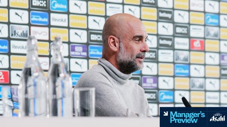 A manager sits at a press conference table before a backdrop featuring multiple logos, including Puma and Etihad, with water bottles and a microphone visible.