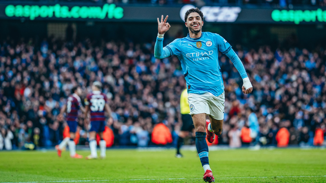 A football player in Manchester City kit celebrates a goal with a crowd in the background.