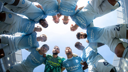 Manchester City team in a huddle wearing light blue jerseys inside Etihad Stadium.