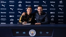 Two people seated at a table with Manchester City branding. One person is signing a document and both are smiling.