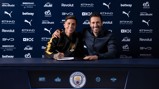 Two people seated at a table with Manchester City branding. One person is signing a document and both are smiling.