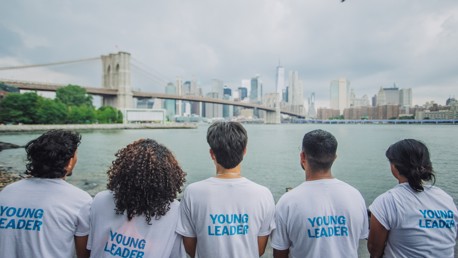 Five people wearing 'Young Leader' shirts sit facing the New York City skyline, with the Brooklyn Bridge in the background.