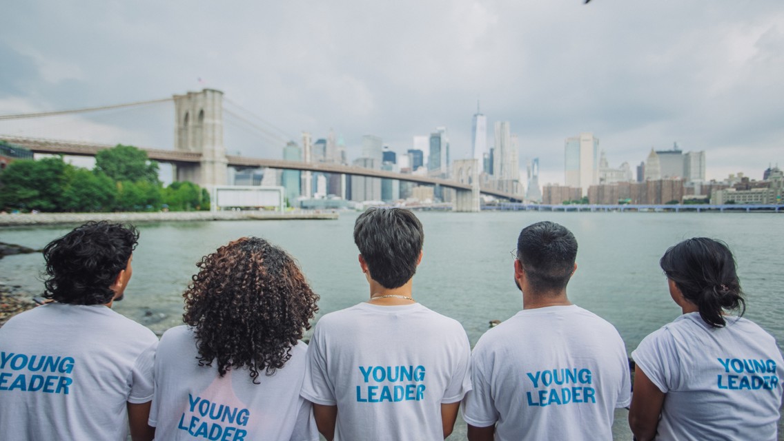 Five people wearing 'Young Leader' shirts sit facing the New York City skyline, with the Brooklyn Bridge in the background.