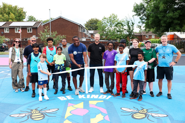 Group of people at a community event standing on a basketball court. Several individuals are holding a ribbon for a ceremony, with the court featuring bee graphics and the phrase 'BE KIND' on the floor.