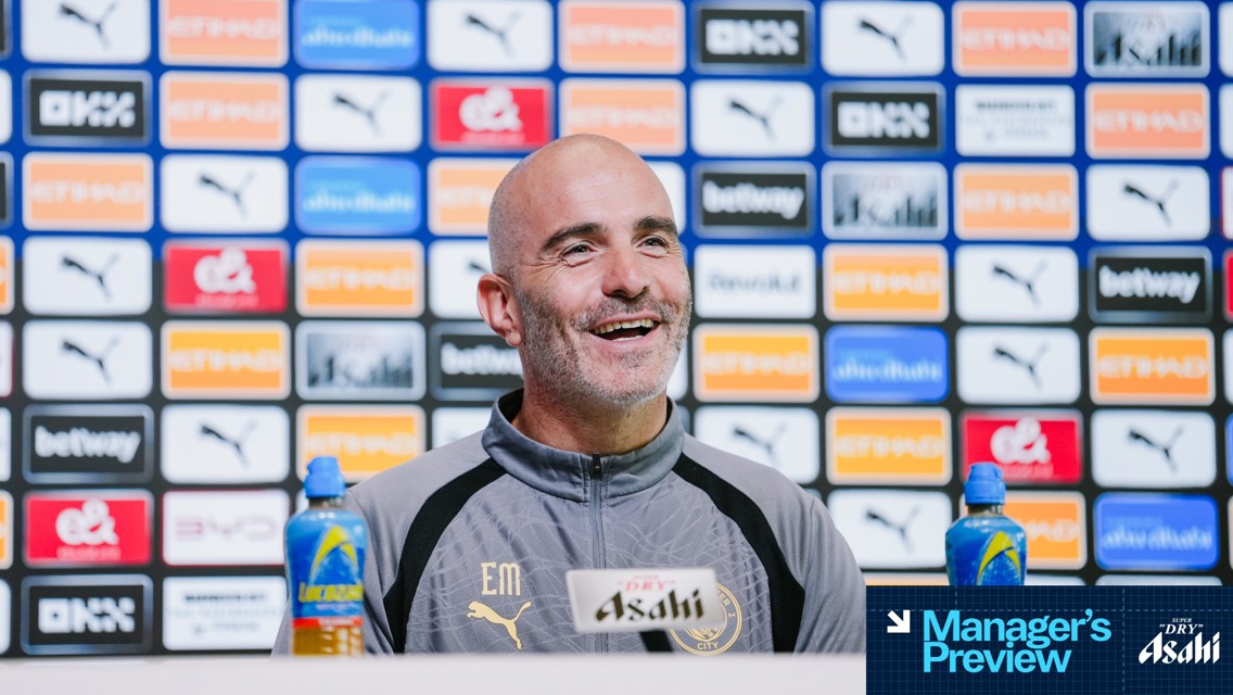 A person speaking at a press conference with a Manchester City logo on the backdrop and table, featuring various sponsor logos.