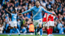 A Manchester City player celebrating a goal, wearing the team's blue kit with arms outstretched. The crowd is visible in the background.