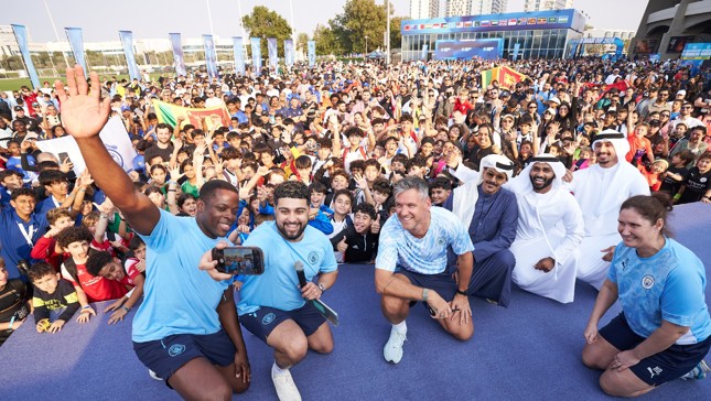A large crowd gathers at an outdoor event with people wearing Manchester City attire and posing for a selfie with a diverse group of attendees.