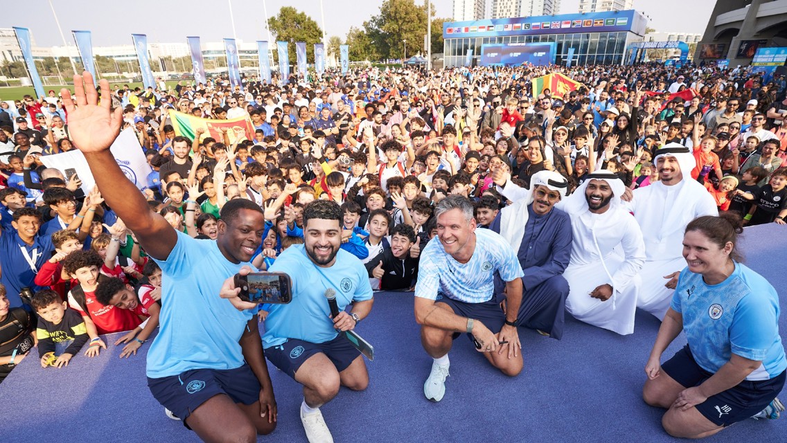 A large crowd gathers at an outdoor event with people wearing Manchester City attire and posing for a selfie with a diverse group of attendees.