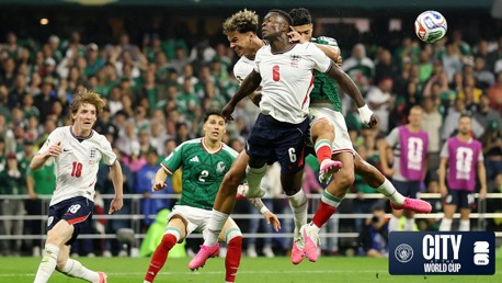 Action shot from a soccer match featuring players from England and Mexico attempting to control the ball mid-air during the World Cup. The crowd is visible in the background.