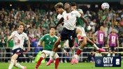 Action shot from a soccer match featuring players from England and Mexico attempting to control the ball mid-air during the World Cup. The crowd is visible in the background.
