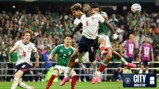 Action shot from a soccer match featuring players from England and Mexico attempting to control the ball mid-air during the World Cup. The crowd is visible in the background.