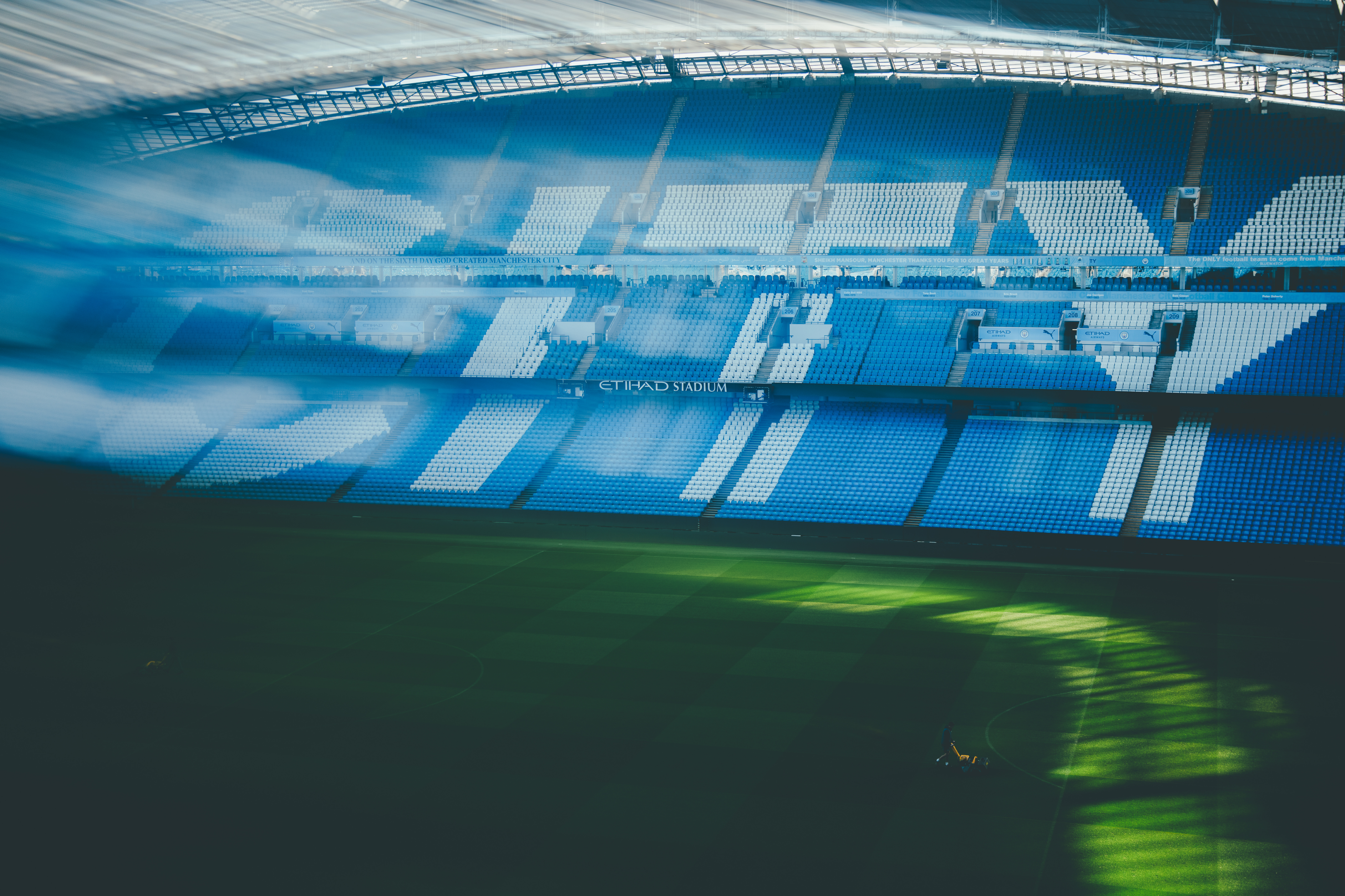 View of the Etihad Stadium with blue seating and sunlight casting shadows on the field.