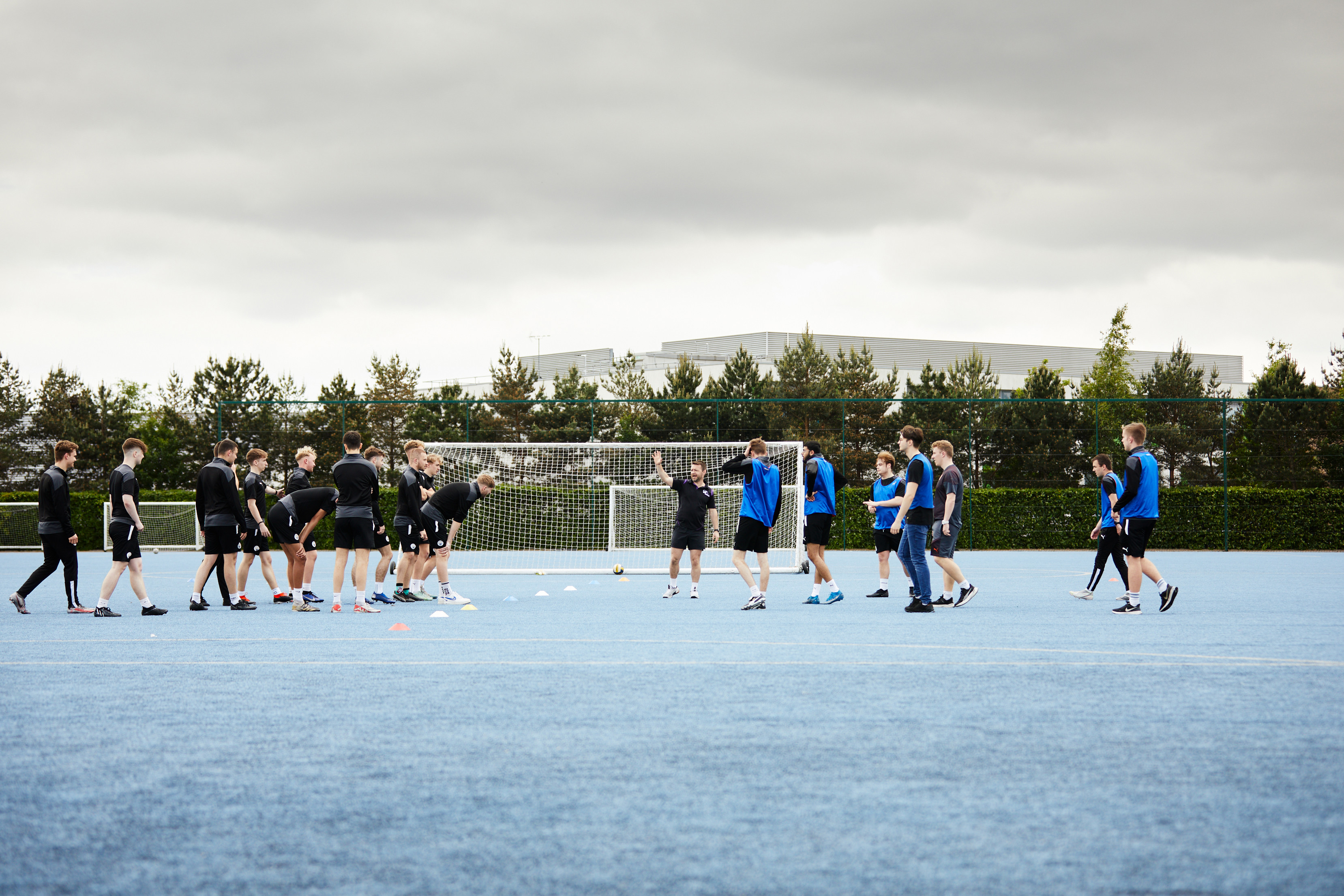 A group of people are training on a blue sports field with cones set up for drills. Some participants are wearing black clothing while others wear blue bibs.