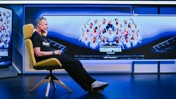 Man sitting in a chair facing a screen showing a women's football team photo with 'Champions 2025/26 Joie Stadium' displayed.
