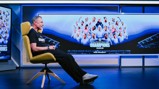 Man sitting in a chair facing a screen showing a women's football team photo with 'Champions 2025/26 Joie Stadium' displayed.