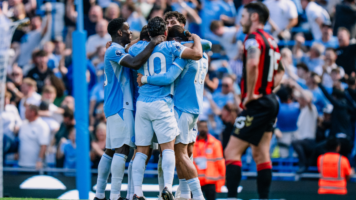 Manchester City players in blue jerseys celebrating a goal with a group hug during a match. An opposing team player in red and black is visible in the foreground. The crowd in the background is cheering.